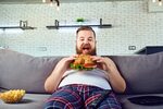 Fat funny man in pajamas eating a burger on the sofa at home