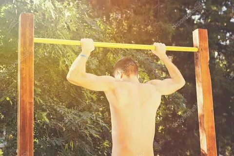 Muscular topless man doing pull-ups on bar outside on sunny 