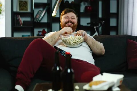 Laughing fat man sits on the sofa eating popcorn and watchin