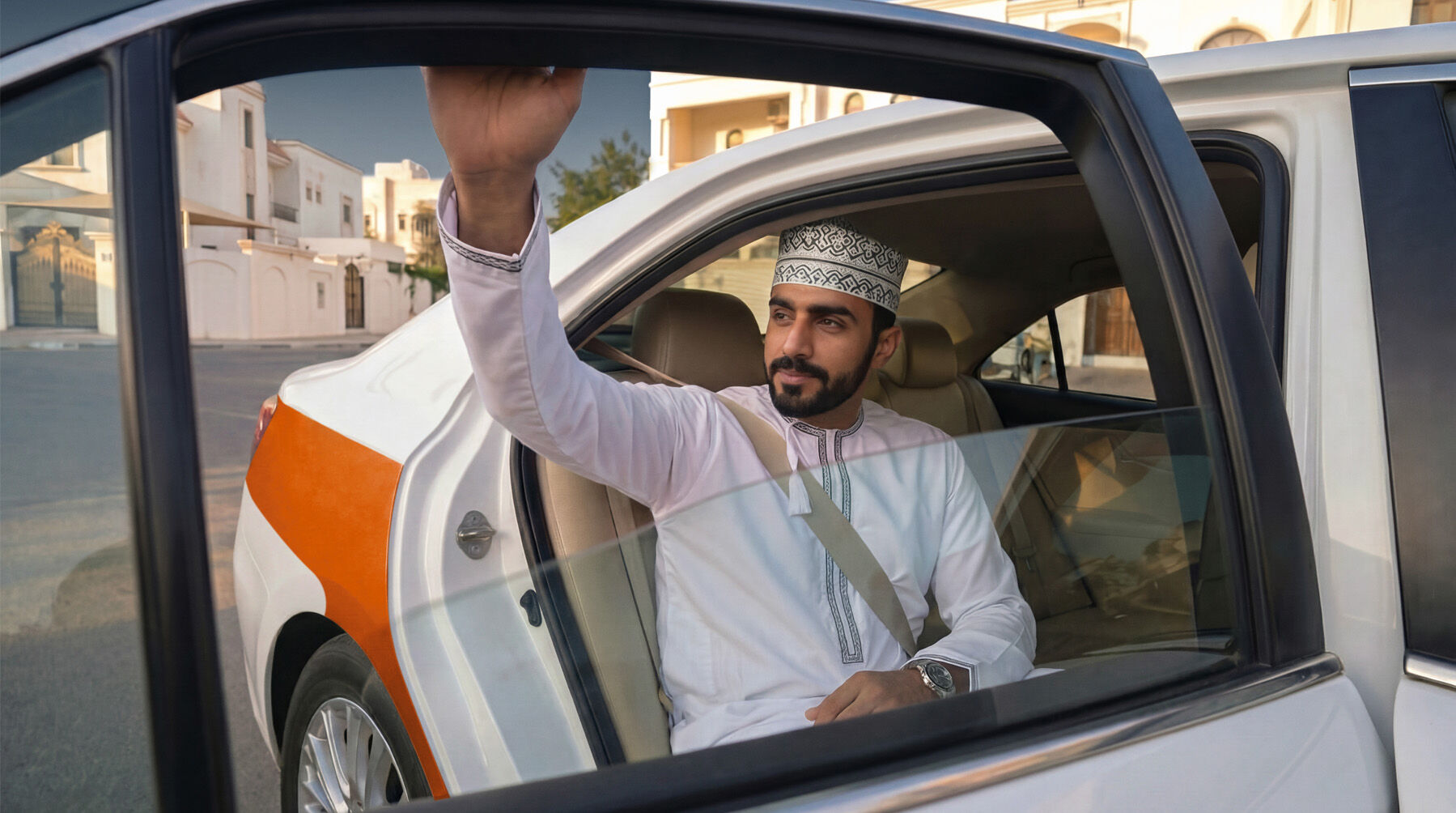 Man in traditional white outfit with patterned headscarf in car, waving with right hand, seatbelt visible, urban background with buildings