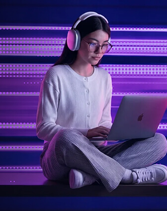 Woman in white sweater, striped pants, white sneakers, wearing headphones and glasses, using Apple laptop