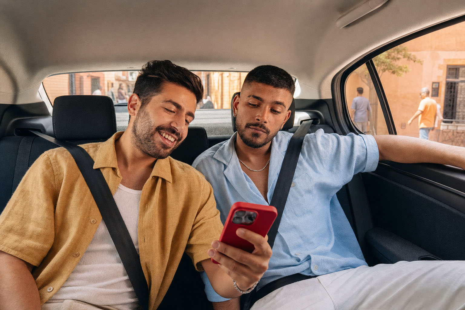 Dos hombres en asientos del auto, uno mira un teléfono rojo, ambos tienen cinturones, ropa casual, uno sonríe, otro mira el teléfono.