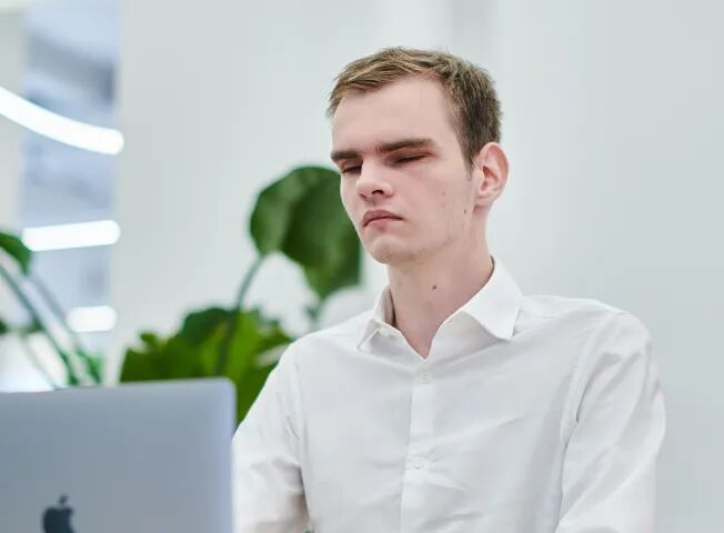 Молодой человек в белой рубашке, работает за ноутбуком Apple, фон — светлый офис с зелёными растениями