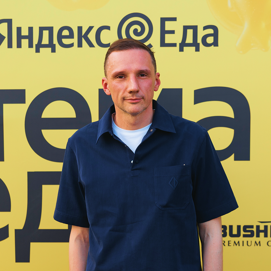 Мужчина в тёмно-синей рубашке на фоне жёлтого баннера с надписью «Яндекс Еда» и частично видимым логотипом Bush