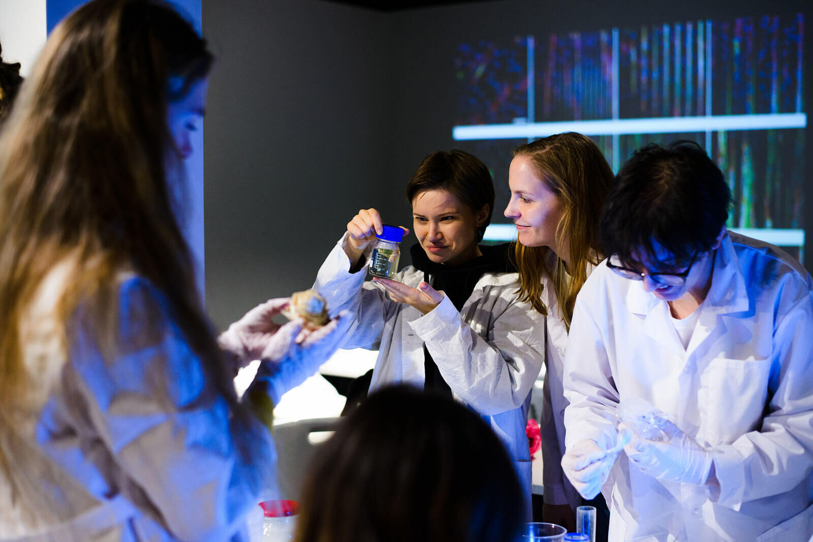 People in lab coats, one holding a jar with blue lid, others handling food and lab equipment