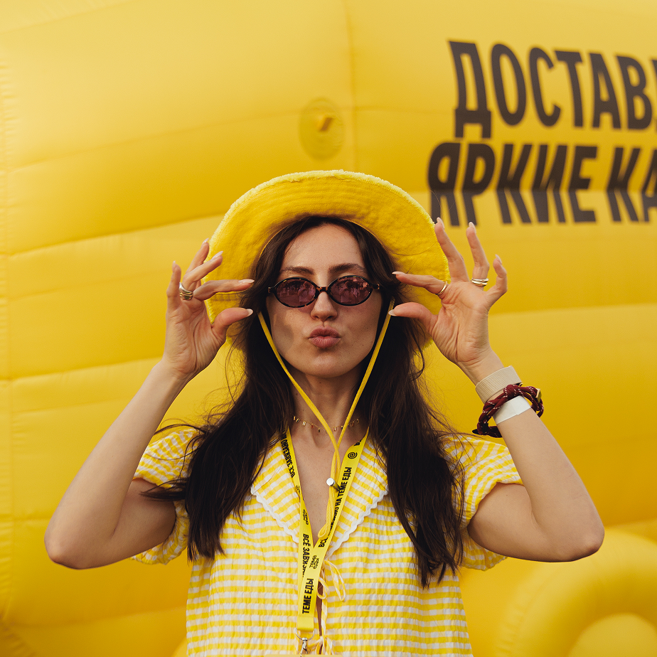 Женщина в жёлтой шляпе и клетчатой одежде, в солнцезащитных очках, на фоне жёлтой стены с надписью «ДОСТАВЛЯЕМ ЯРКИЕ КА…».