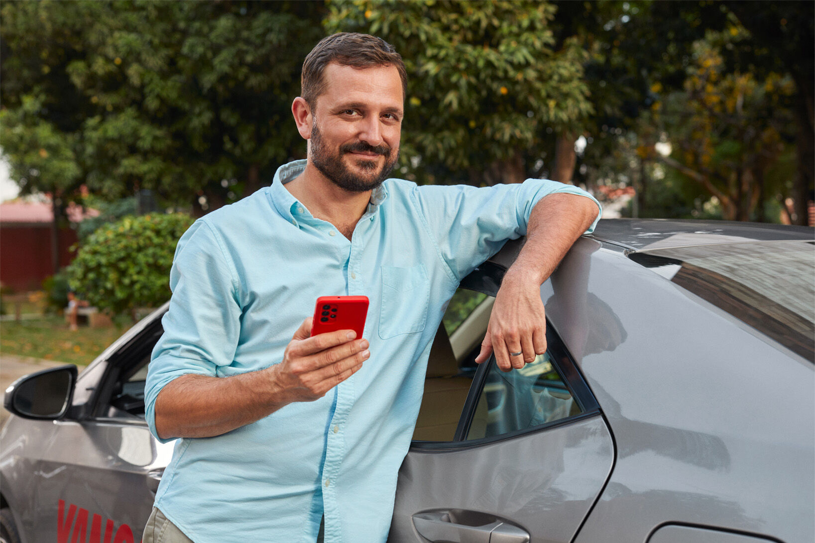 Man in light blue shirt leaning on car, holding red smartphone