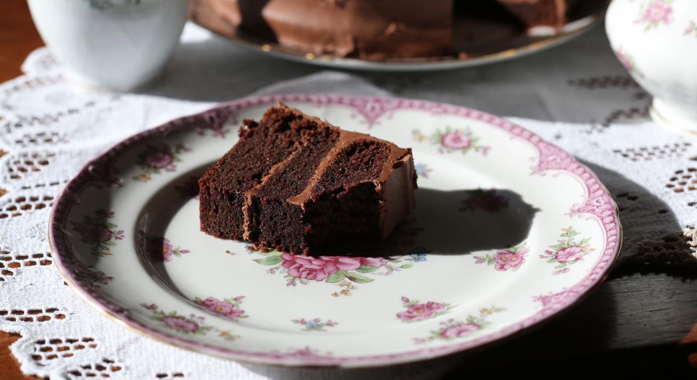 A delicate porcelain dessert plate with intricate floral patterns, holding a slice of rich chocolate cake, placed on a lace-lined wooden table in a sunlit Turkish kitchen.