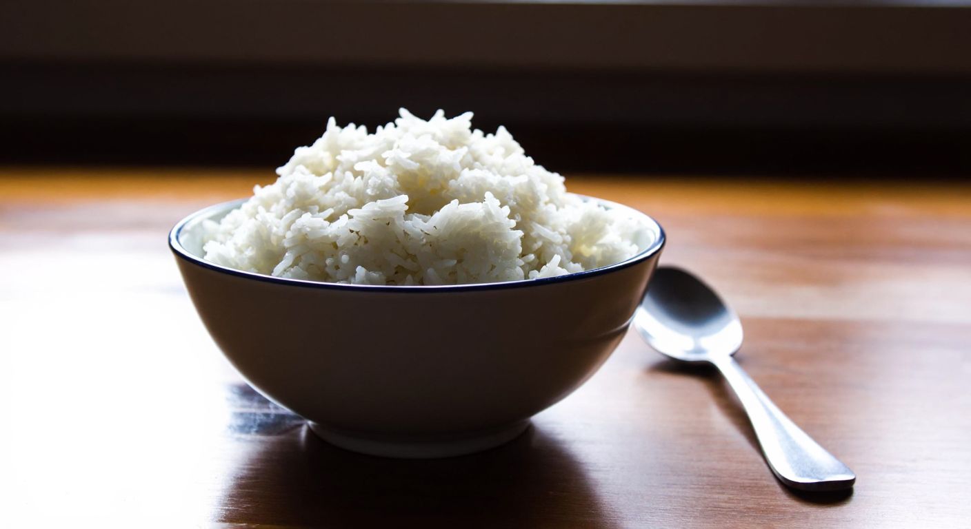 A small bowl of fluffy white rice sits on a wooden table in a Turkish kitchen, with a spoon resting beside it, conveying simplicity and naturalness.