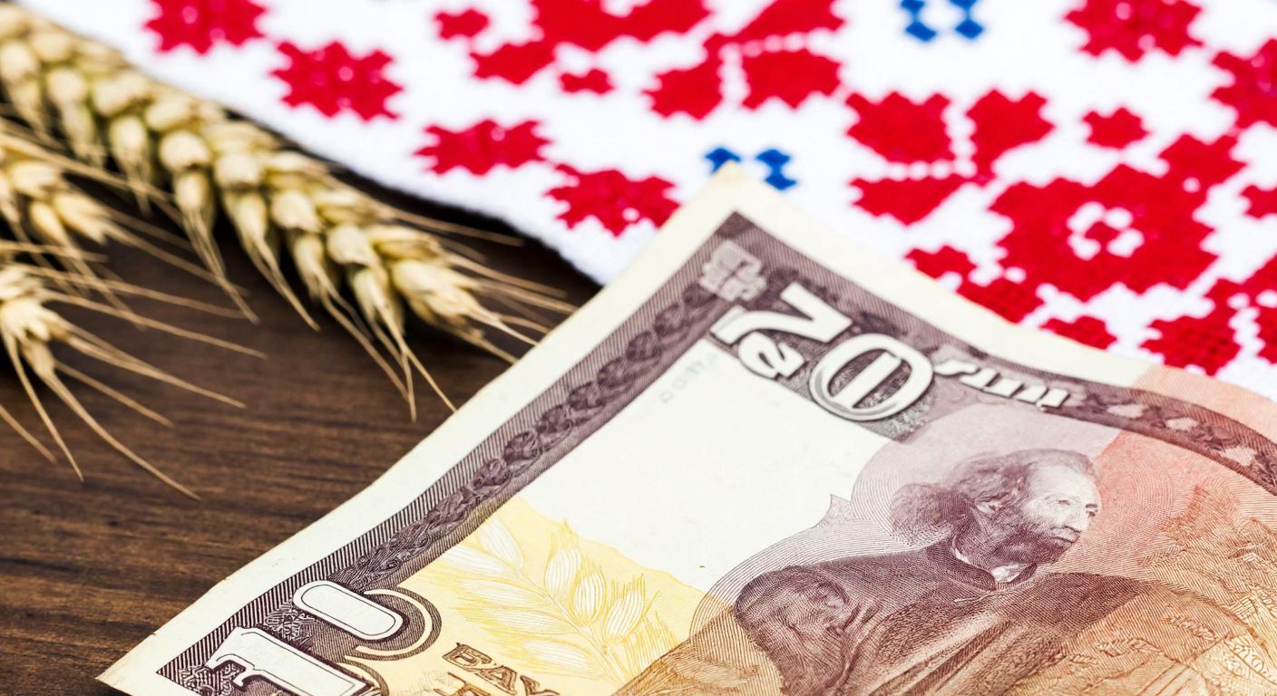 A close-up of a weathered Ukrainian grivna banknote resting on a wooden table, with golden wheat fields and traditional embroidery patterns faintly visible in the background.