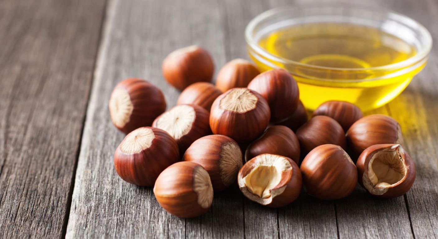 A close-up of a small pile of golden-brown hazelnuts on a rustic wooden table, with a few cracked open to reveal their creamy interiors, next to a small glass bowl filled with rich, golden hazelnut oil.