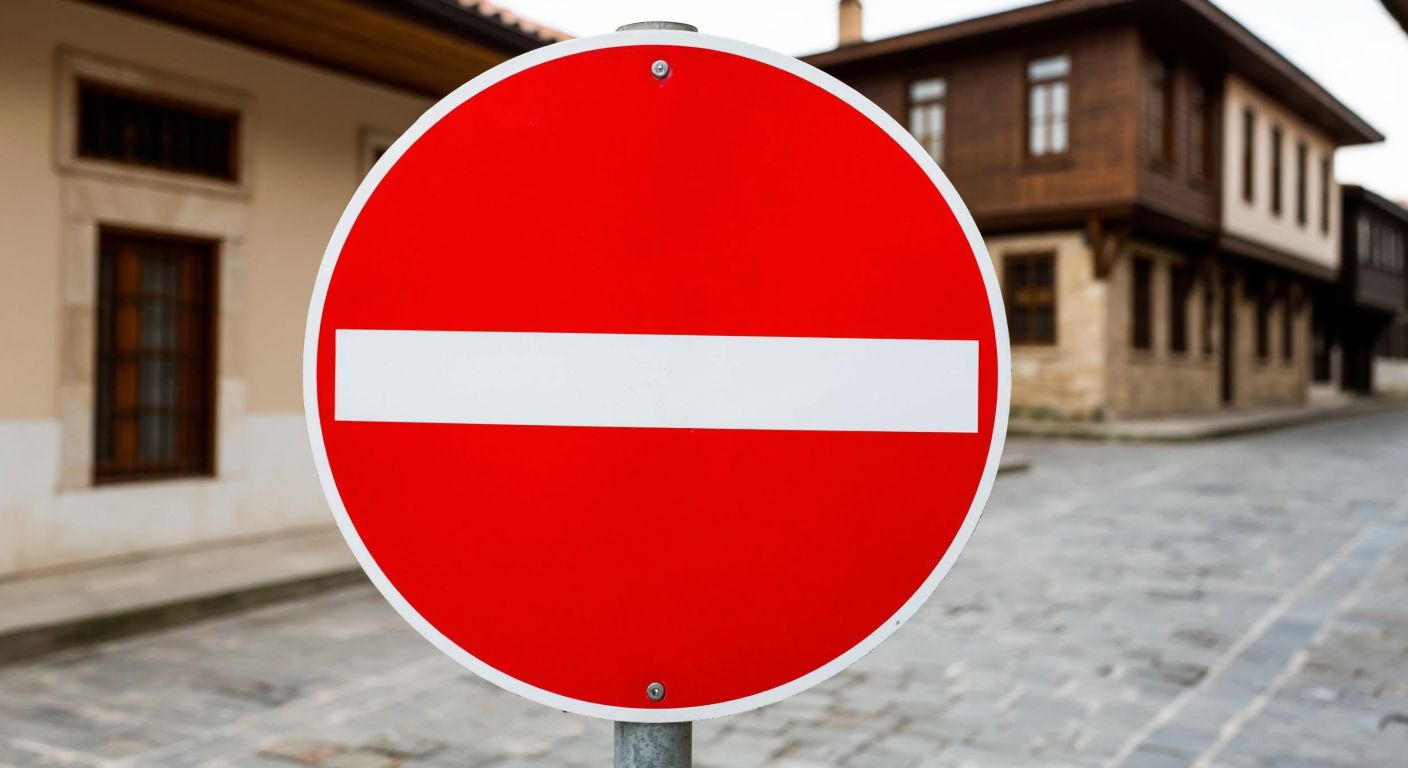 A red circular road sign with a white horizontal bar, resembling a "No Entry" symbol, placed against a blurred background of a Turkish street with cobblestones and traditional Ottoman-style buildings.