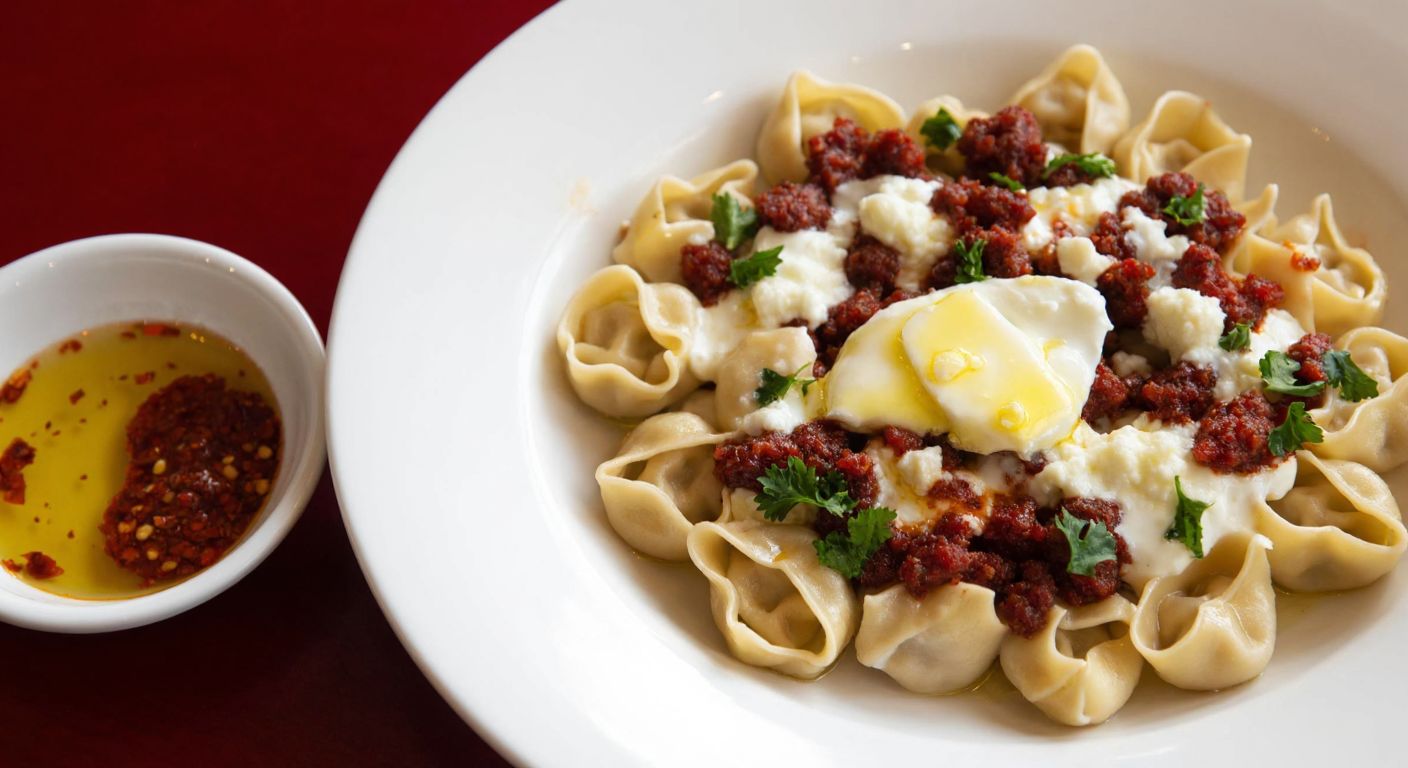 A steaming plate of Turkish manti topped with garlic yogurt and melted butter, with a small bowl of red pepper oil beside it, evoking warmth and comfort.