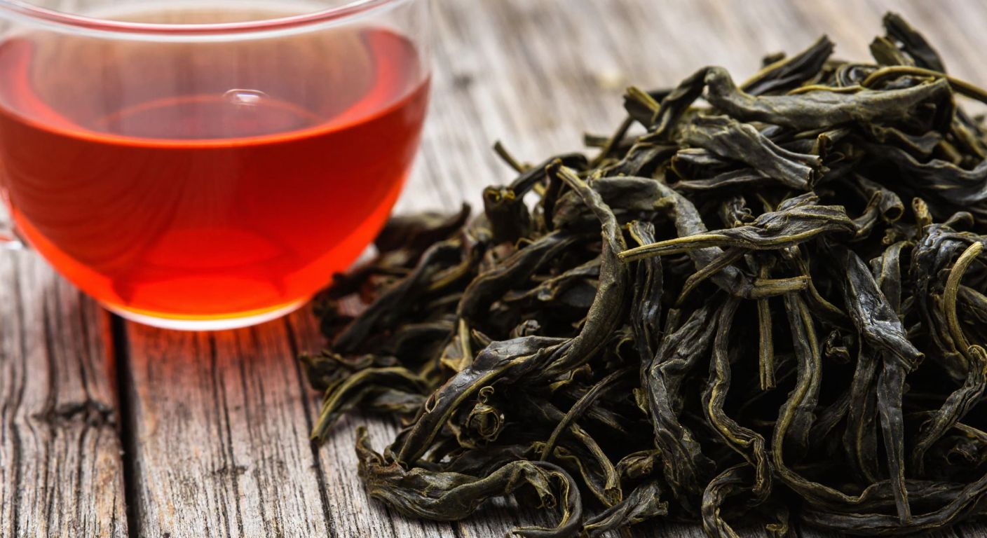 A close-up of vibrant, dark green tea leaves with a glossy sheen, neatly curled and uniform in size, resting on a rustic wooden surface next to a glass of freshly brewed tea glowing with a bright copper-red hue.