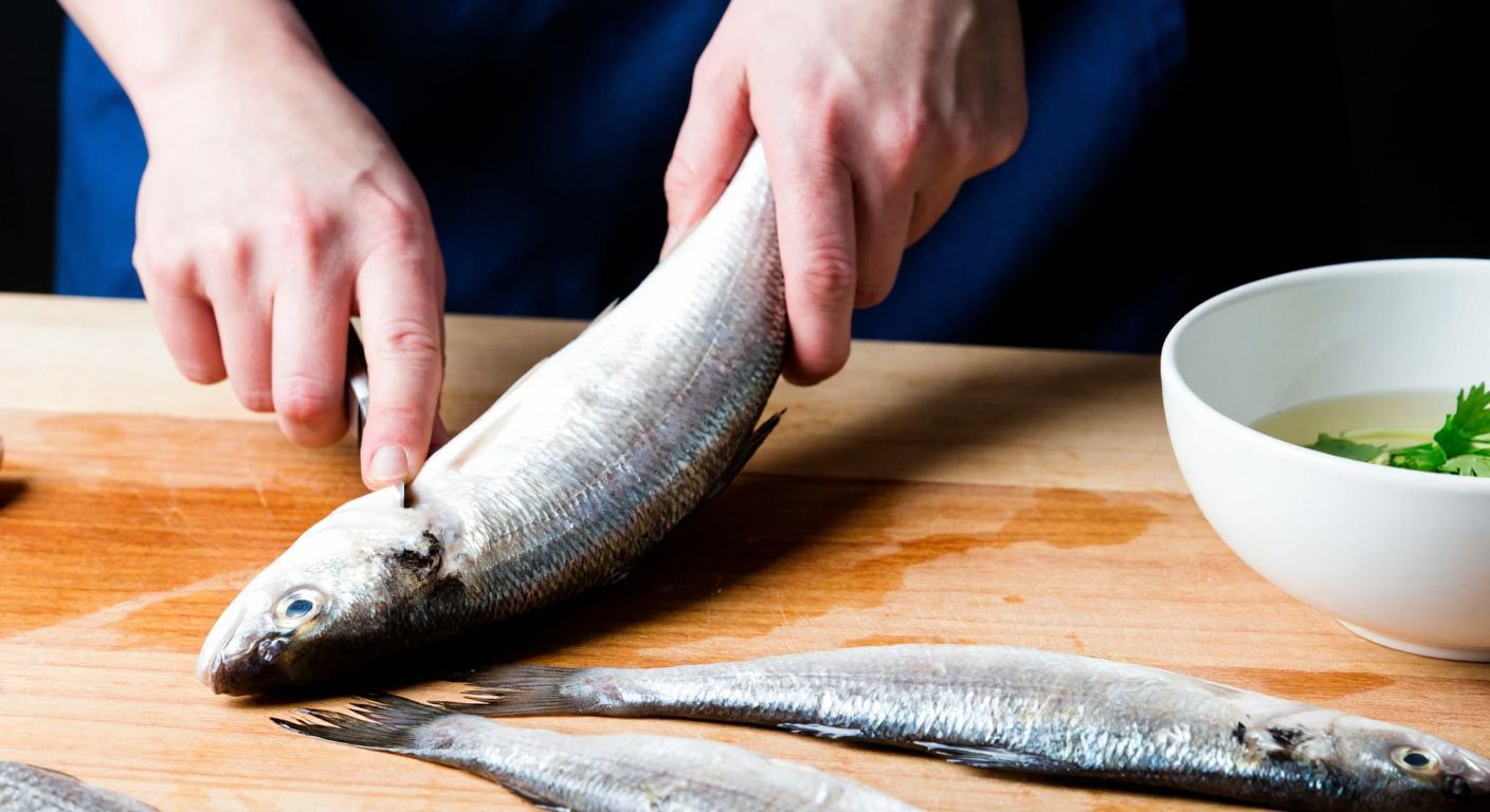 A close-up of a skilled cook’s hands using a sharp knife to carefully fillet a fresh whiting fish on a wooden cutting board, with a bowl of water nearby and scattered fish scales glistening under warm kitchen light.