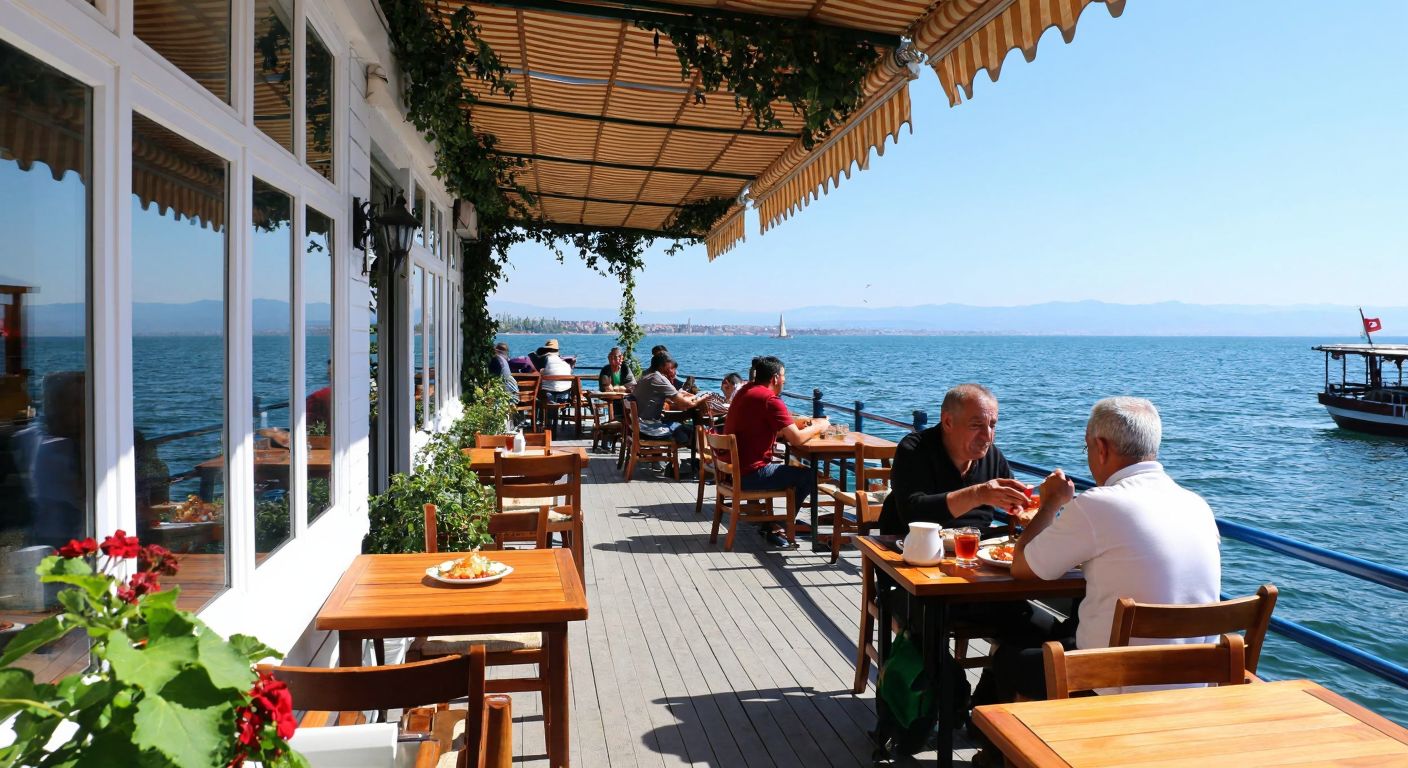 A cozy waterfront café in İzmit with white wooden décor, patrons enjoying tea and traditional Turkish dishes like menemen, and a serene view of the sea under a clear blue sky.