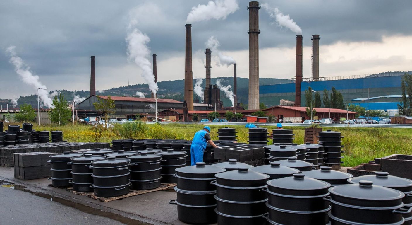 A sprawling industrial factory in Kocaeli with towering chimneys emitting steam, surrounded by stacks of cast iron pots and enamel-coated cookware under a cloudy sky, with workers in blue overalls inspecting finished products.