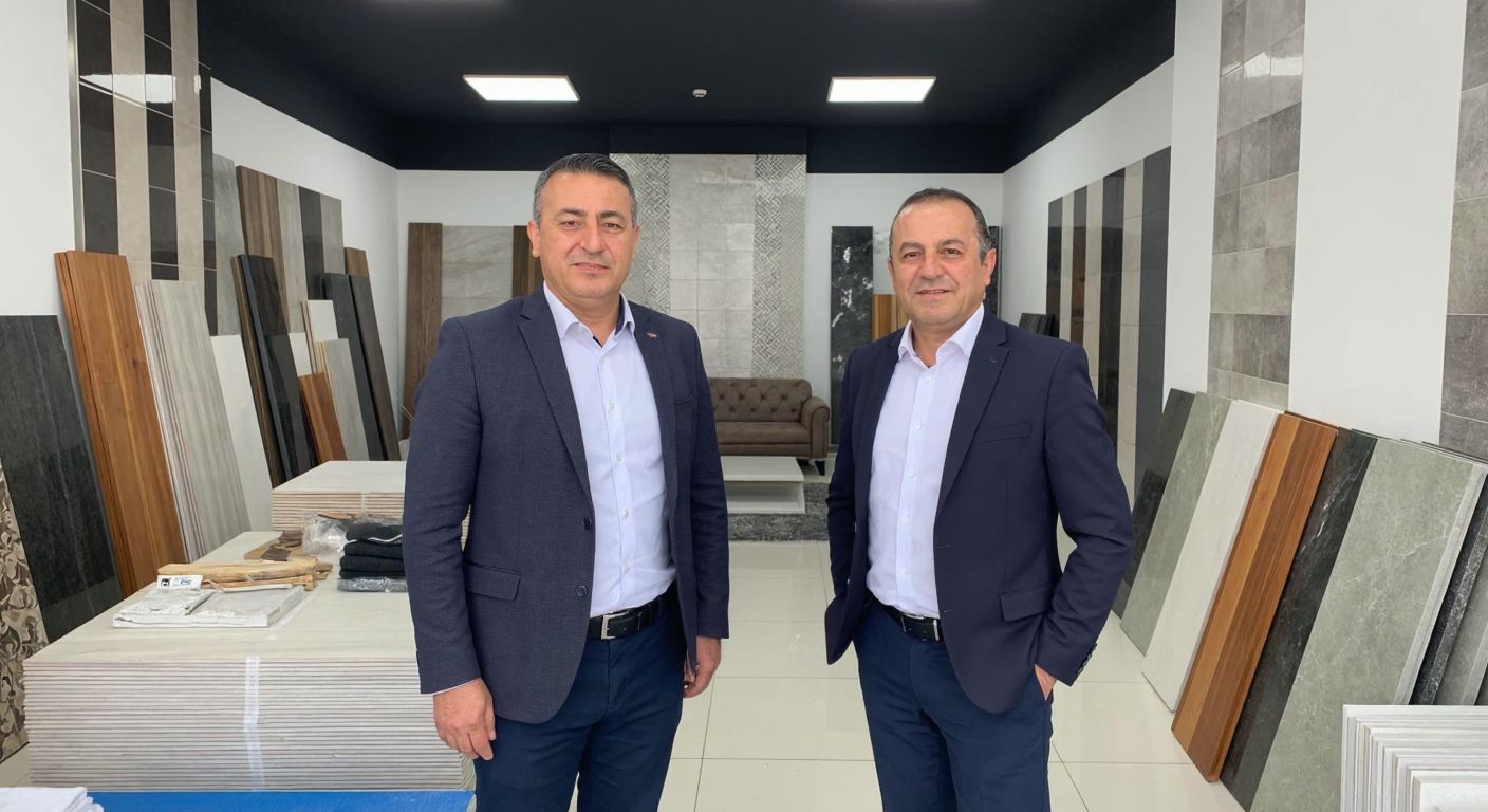 Two middle-aged Turkish men in business attire stand proudly in front of a ceramic tile showroom in İzmit, surrounded by colorful tile displays and construction materials.