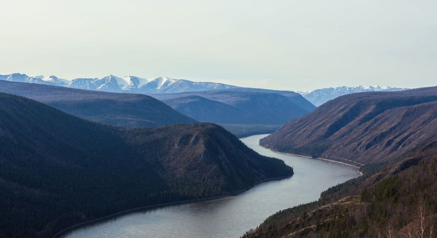 A vast, winding river (Yenisey) cuts through a rugged Siberian landscape, flanked by dense forests and distant snow-capped mountains under a pale blue sky.