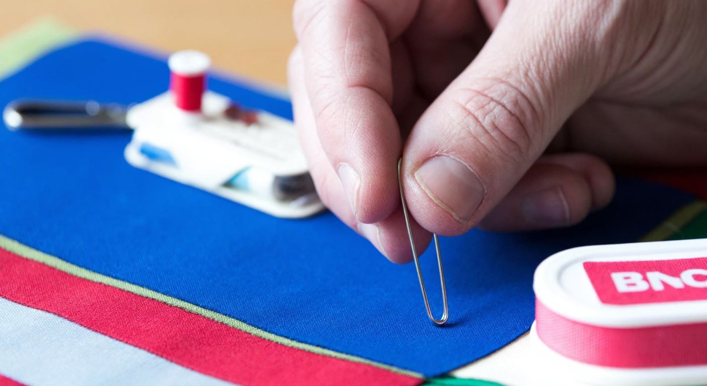 A close-up of a weathered hand holding a safety pin against a colorful patch of fabric, with a small sewing kit and a first-aid bandage visible in the blurred background.