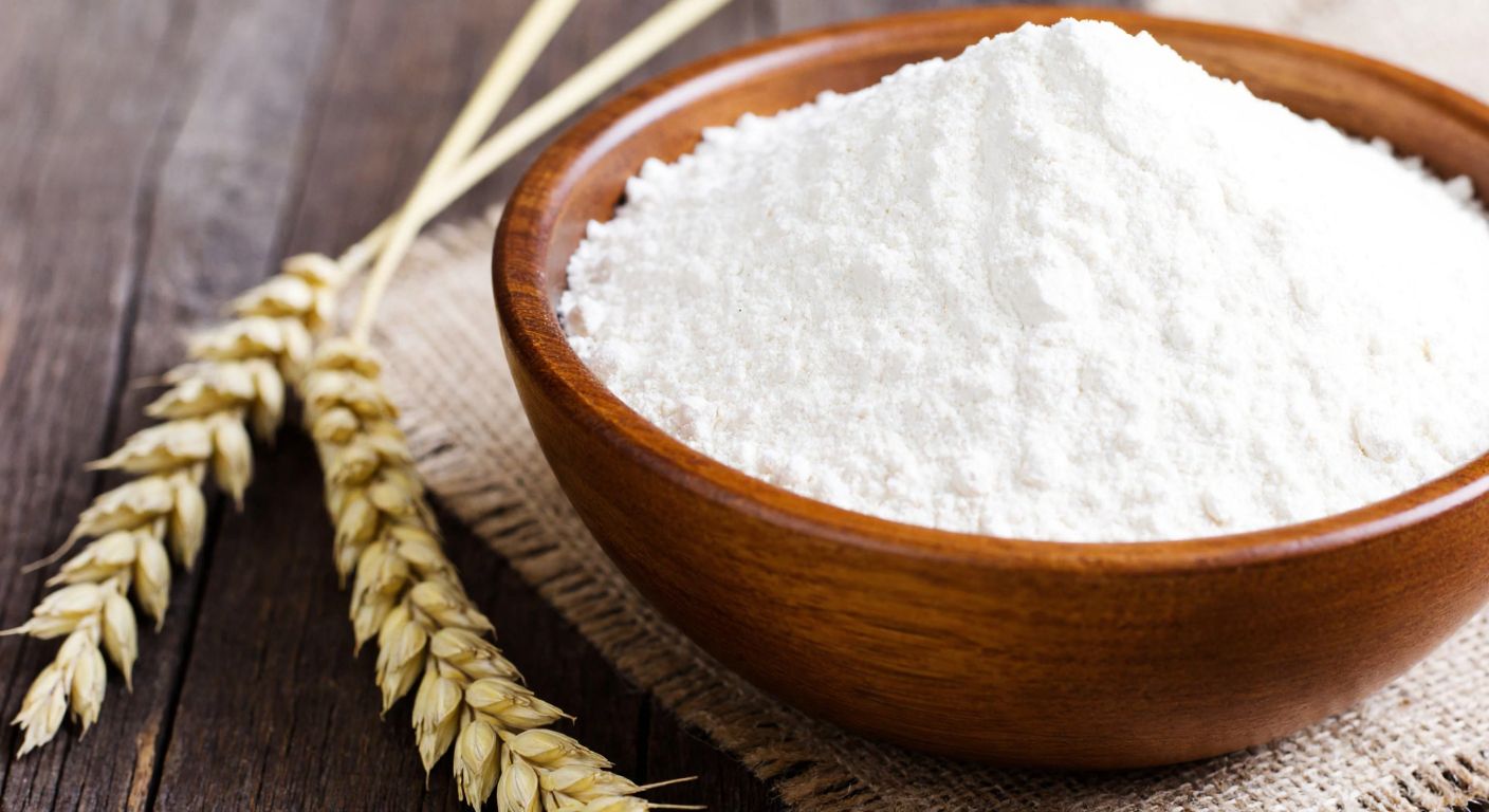 A wooden bowl filled with fine white flour sits on a rustic table next to a golden wheat stalk, evoking simplicity and nourishment.