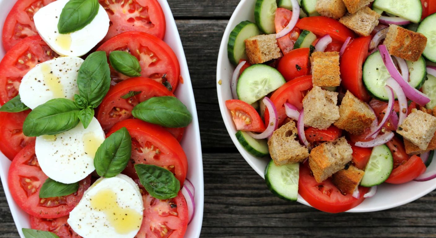 A rustic wooden table holds two vibrant salads side by side: one with sliced red tomatoes, creamy mozzarella, and fresh green basil leaves drizzled with golden olive oil, and the other with chunky tomatoes, cucumbers, onions, and crusty bread cubes tossed in a glossy vinaigrette.