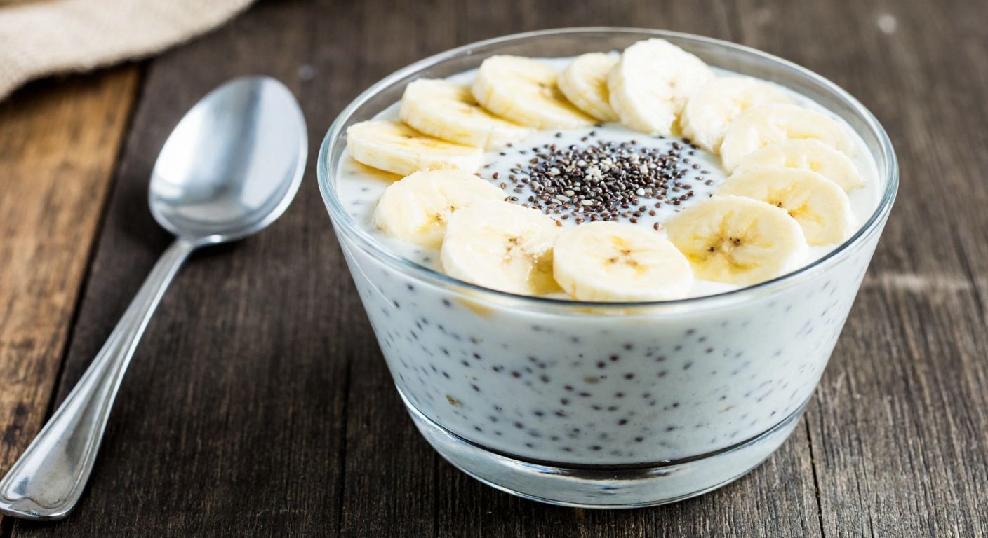 A creamy white yogurt pudding in a glass bowl, topped with chia seeds and sliced bananas, placed on a rustic wooden table with a spoon beside it, evoking freshness and wholesome nutrition.
