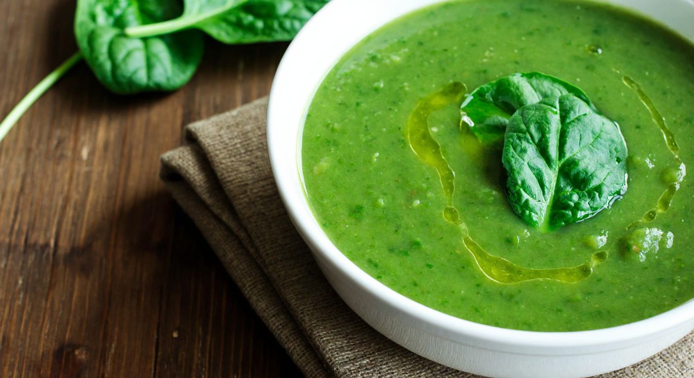 A steaming bowl of vibrant green spinach soup sits on a rustic wooden table, with fresh spinach leaves and a drizzle of olive oil visible on top, evoking warmth and simplicity.