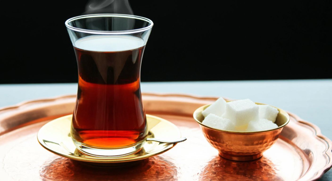 A delicate, tulip-shaped Turkish tea glass with a narrow waist, filled with steaming amber tea, resting on a small saucer with a golden rim, placed on a copper tray beside a sugar cube bowl.