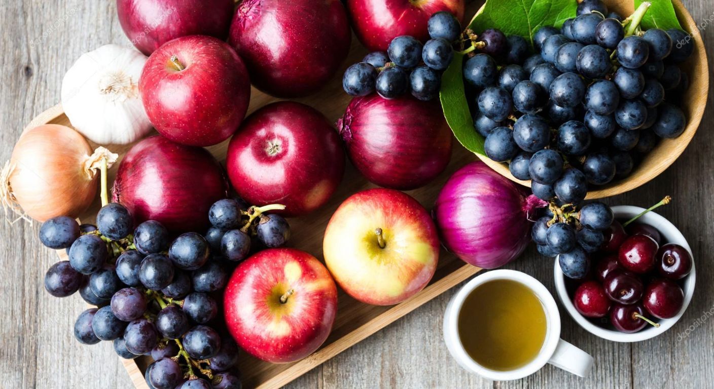 A vibrant still life of fresh red apples, purple onions, and bunches of dark grapes arranged on a rustic wooden table, with a small bowl of cherries and a steaming cup of green tea nearby.