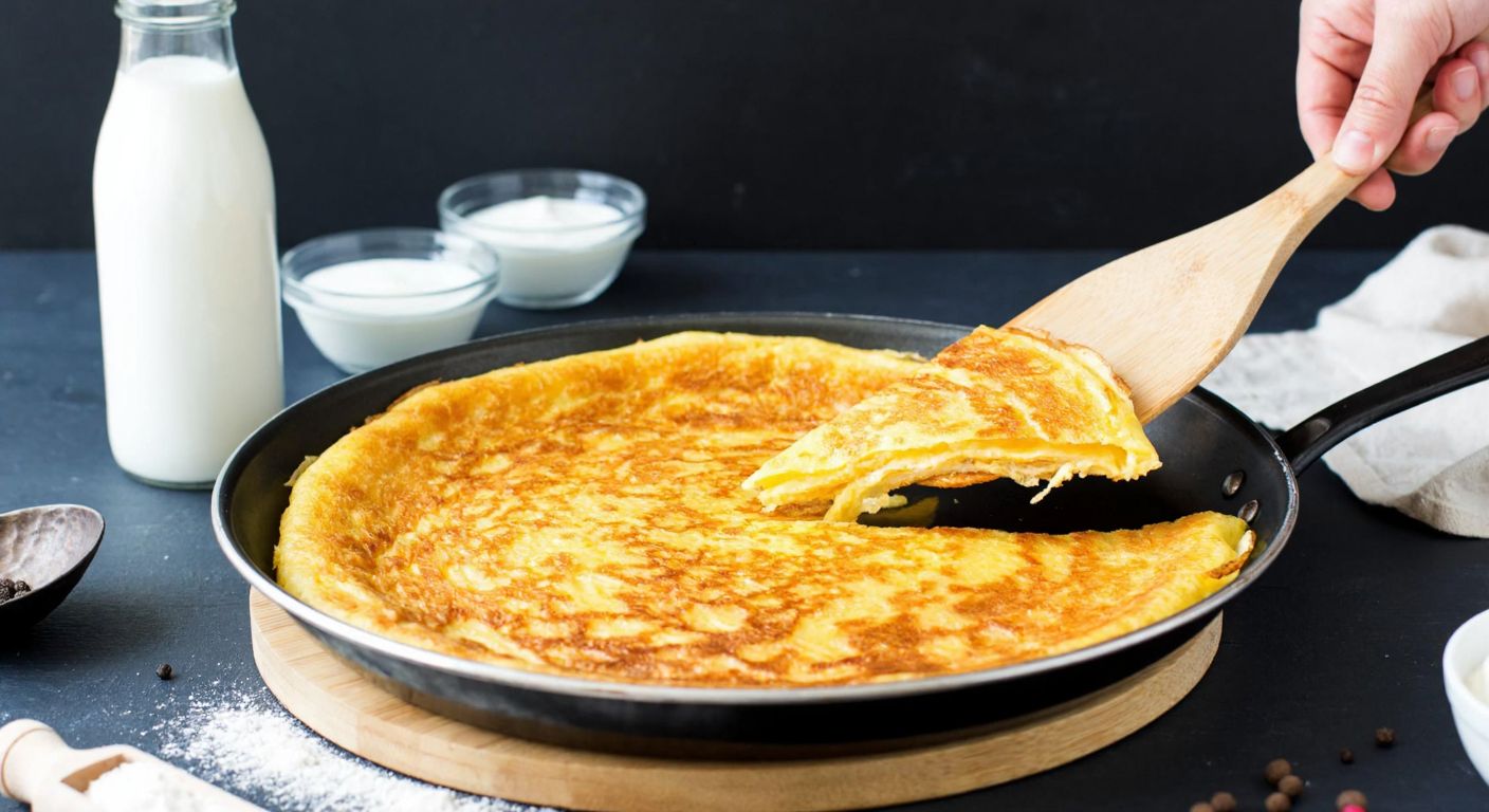 A warm, golden-brown eggless crepe sizzling in a traditional Turkish pan, with a wooden spatula lifting its crispy edge, surrounded by fresh ingredients like milk, yogurt, and flour on a rustic kitchen counter.