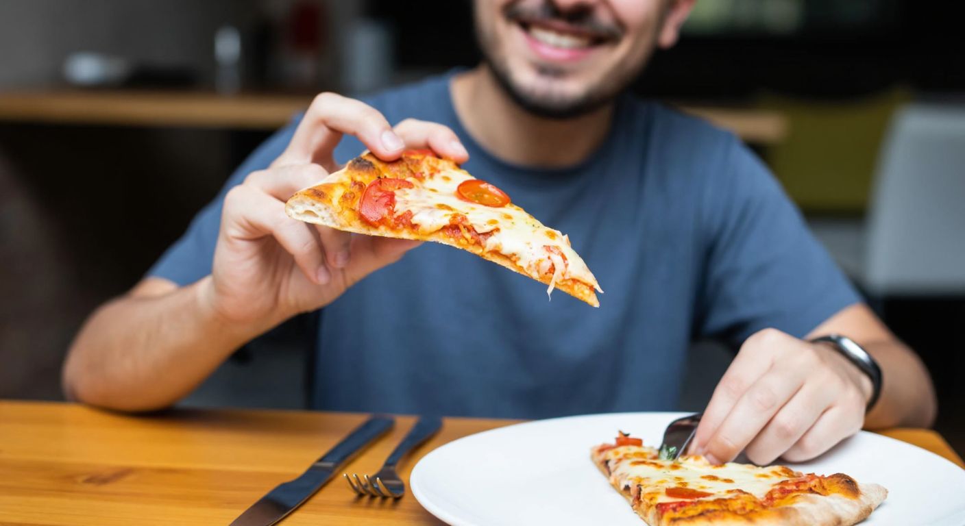 A person in a casual Turkish home setting holds a slice of thin-crust pizza with their fingers, smiling as they take a bite, with a fork and knife resting unused on a wooden table nearby.