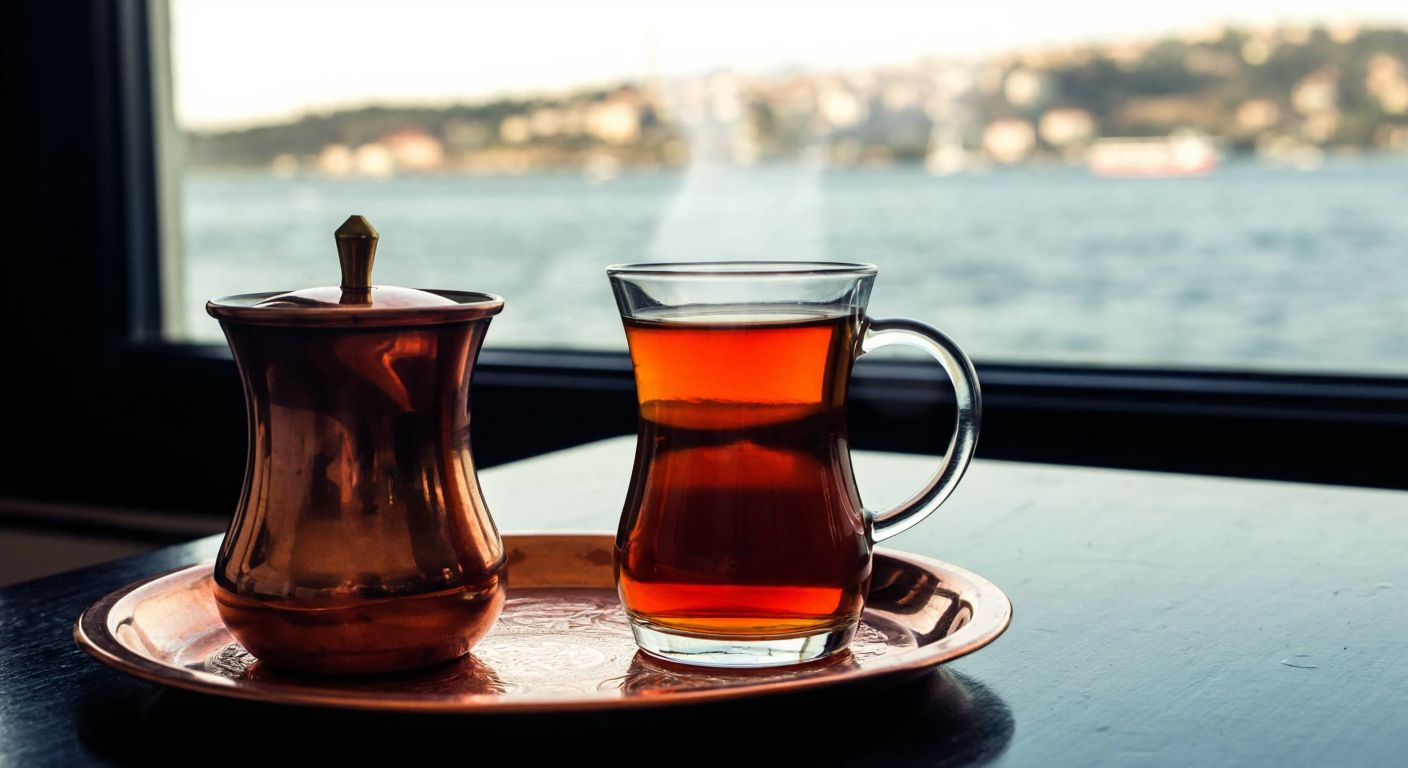 A warm, steaming glass of amber-colored tea sits on a small copper tray beside a traditional Turkish tea glass, with a serene Bosphorus view in the background.