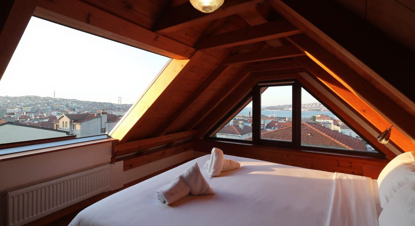 A cozy attic room with slanted wooden beams, bathed in warm sunlight streaming through a large, open skylight, overlooking a view of Istanbul's rooftops and the Bosphorus.