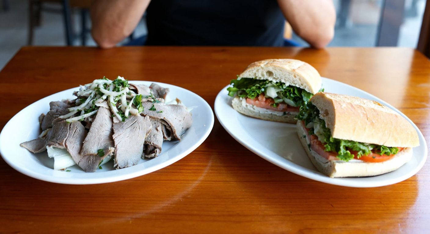 A rustic wooden table in a Turkish eatery holds a plate of thinly sliced, cold boiled lamb offal (söğüş) next to a freshly cut sandwich with simple fillings, evoking curiosity and mild confusion.