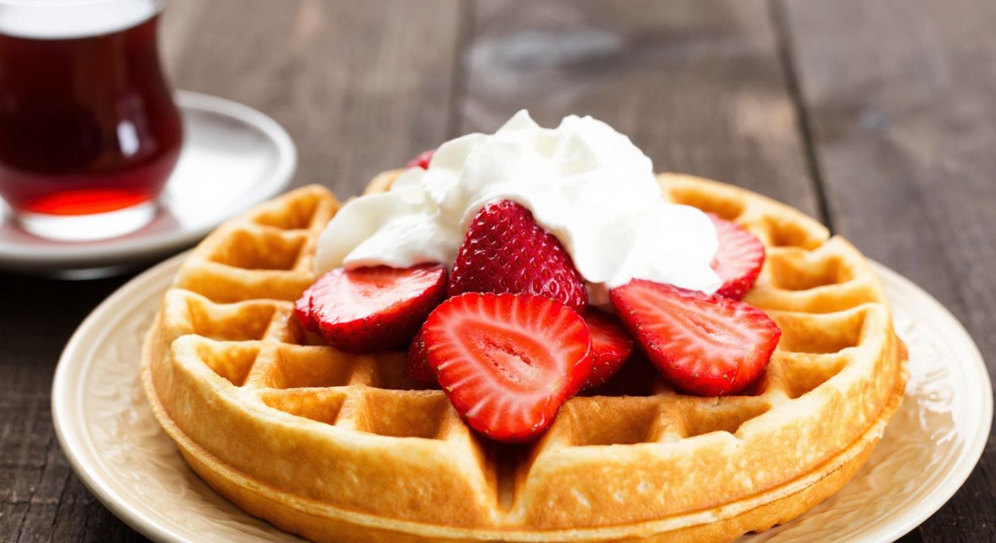 A golden-brown waffle topped with fresh strawberries and whipped cream, placed on a rustic wooden table with a steaming cup of Turkish tea in the background, evoking a cozy, anytime indulgence.