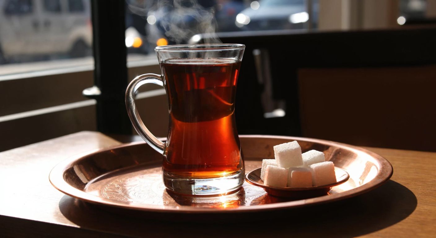 A steaming glass of amber Turkish tea with sugar cubes resting beside it on a small saucer, set against a traditional copper tray in a cozy, sunlit Turkish café.