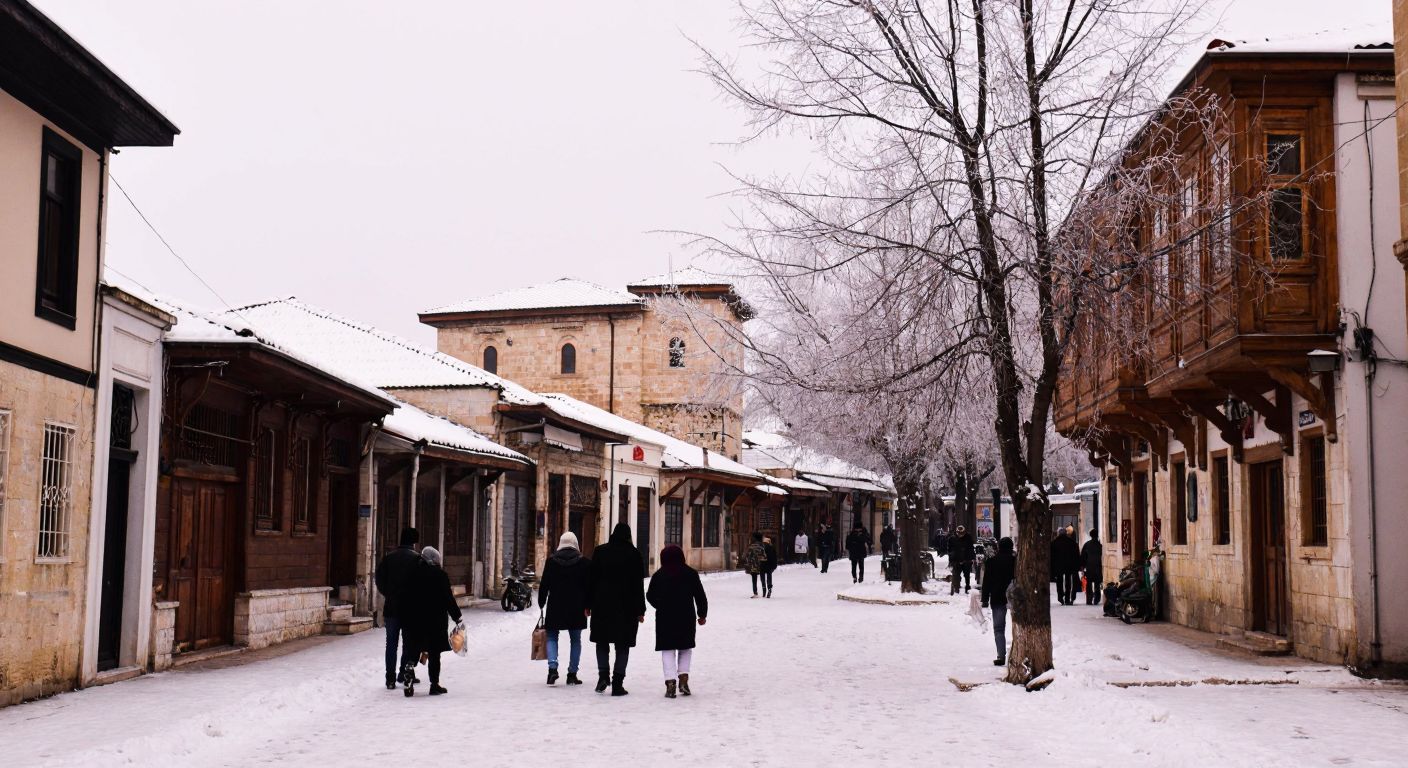 A snowy winter scene in Gaziantep with frost-covered rooftops, bare trees, and bundled-up locals walking through a quiet, cobblestone street under a pale gray sky.
