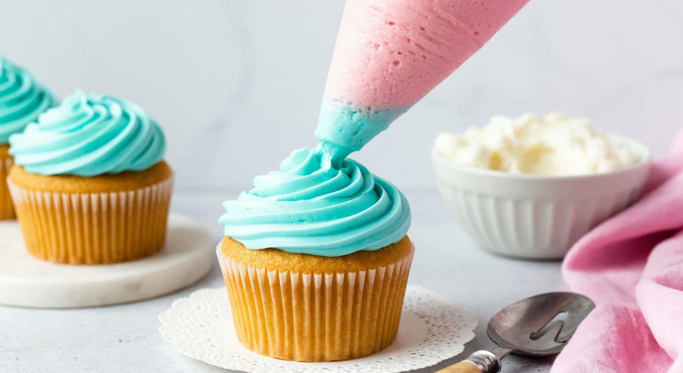 A vibrant, pastel-colored American buttercream frosting being smoothly spread over a golden-brown cupcake, with a small silver spatula and a bowl of fluffy frosting nearby, evoking a sweet and cheerful baking scene.