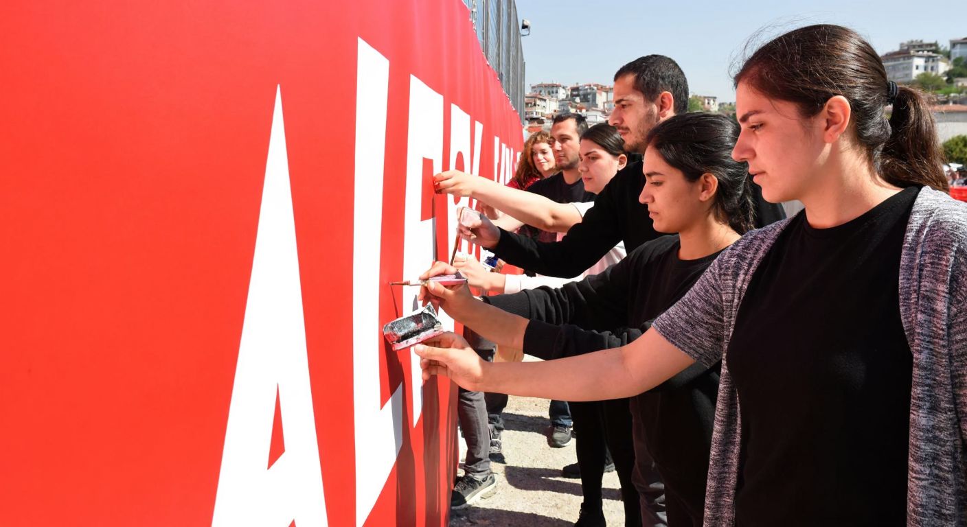 A group of people in Turkey collaboratively painting a large, bright red banner with bold white letters using stencils, their faces focused and determined.