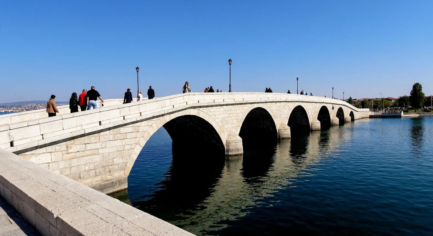 A serene stone bridge with four arched sections stretches across a calm lake in Büyükçekmece, with people strolling along its walkway under a clear blue sky.