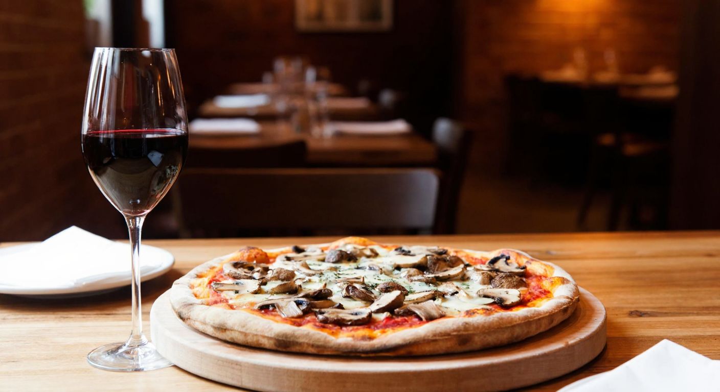 A rustic wooden table in a cozy setting holds a steaming pizza topped with mushrooms and cheese, accompanied by a glass of deep red Pinot Noir, with a dimly lit Italian-style trattoria in the background.