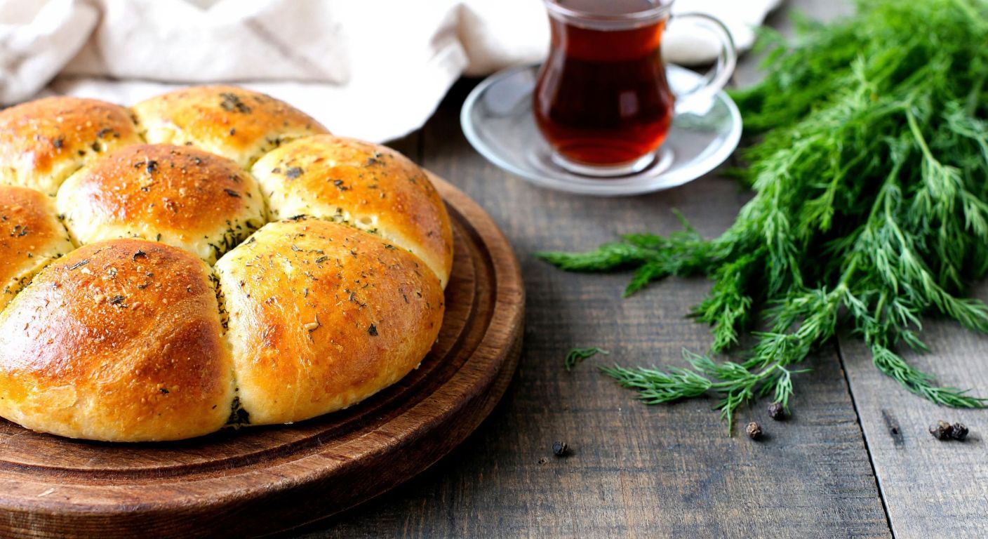 A warm, golden-brown dill-studded poğaça sits on a rustic wooden table beside a small cup of Turkish tea, with fresh dill sprigs scattered nearby, evoking a cozy Turkish home kitchen.