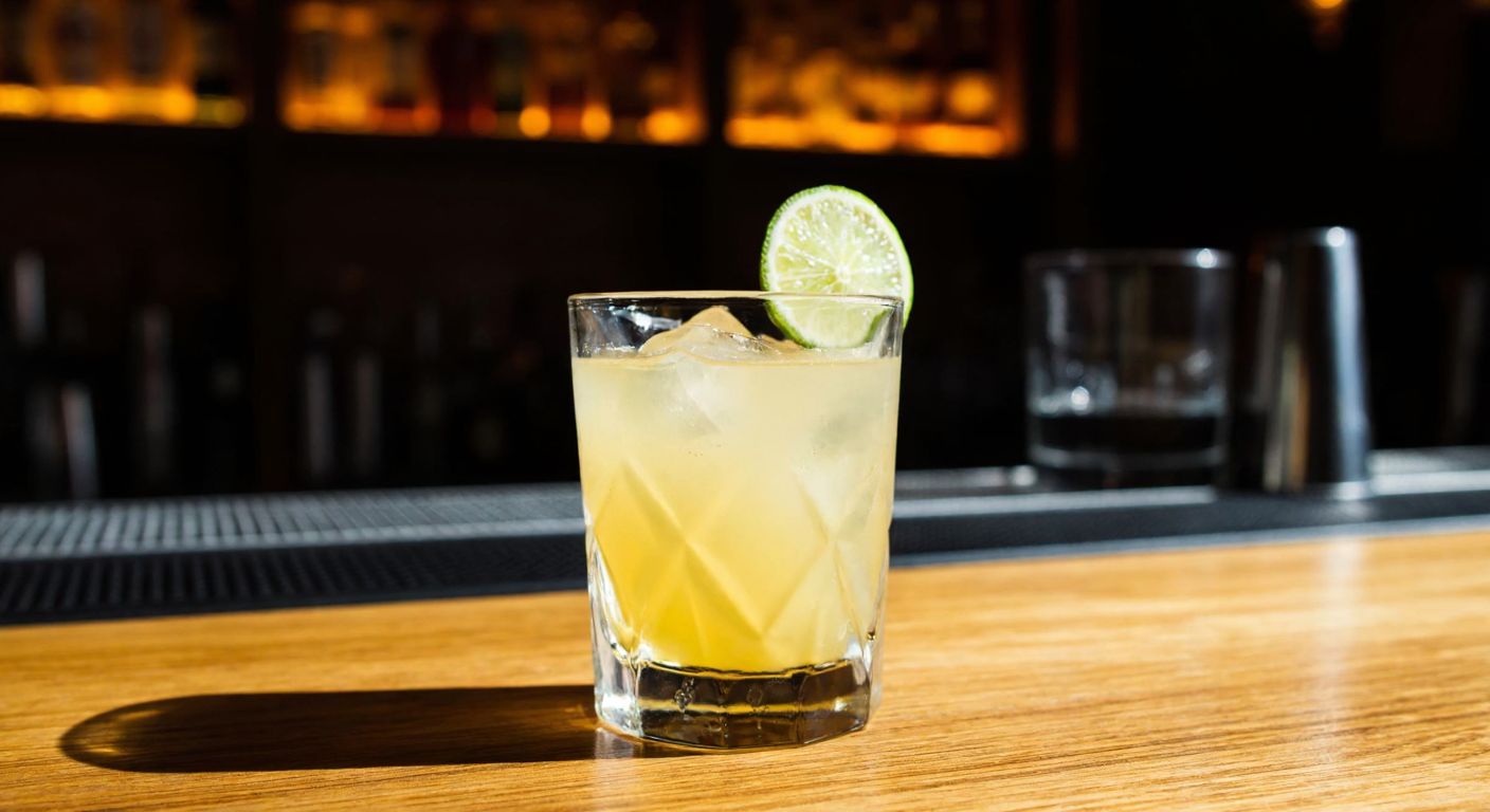 A clear glass filled with golden gin, resting on a wooden bar counter with a slice of lime on the rim, surrounded by dim ambient lighting.