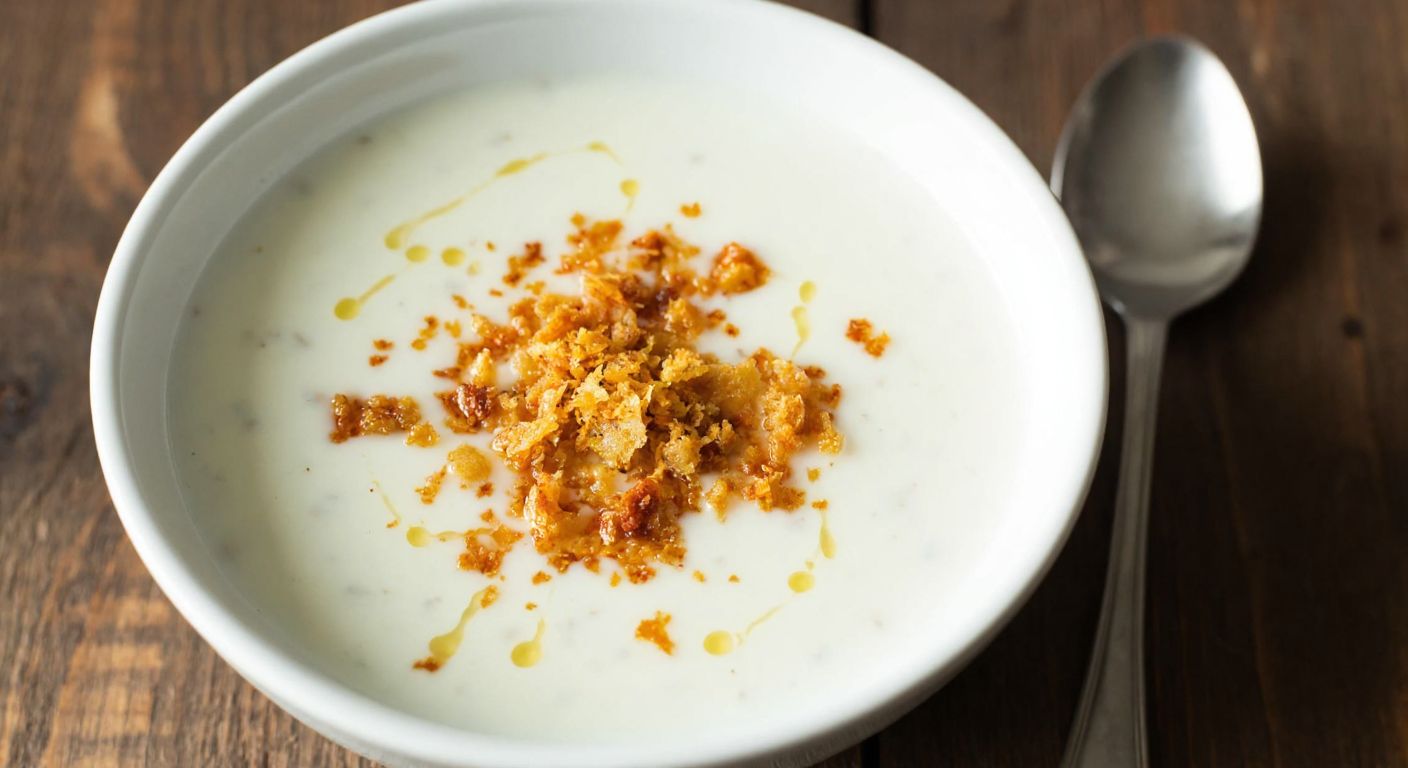 A steaming bowl of creamy yogurt soup with golden tarhana flakes sprinkled on top, set on a rustic wooden table with a spoon beside it, evoking warmth and traditional Turkish comfort.