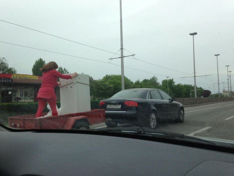 Женщина стоя в прицепе держит холодильник, на полном ходу машины