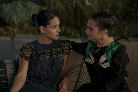 Alicia Vikander and Adria Arjona sit together on a couch. 