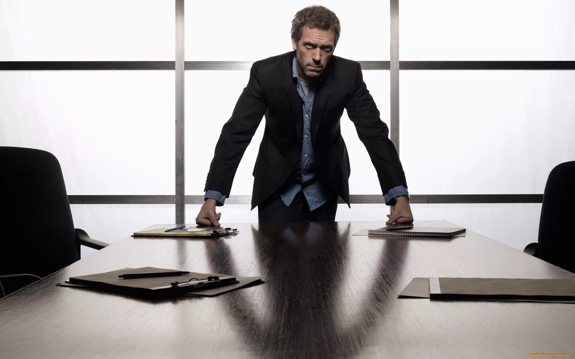 Возле стола представлявшего сильно уменьшенную. Человек опирается на стол. Бизнесмен за столом. Мужчина в костюме в офисе. Властный человек.