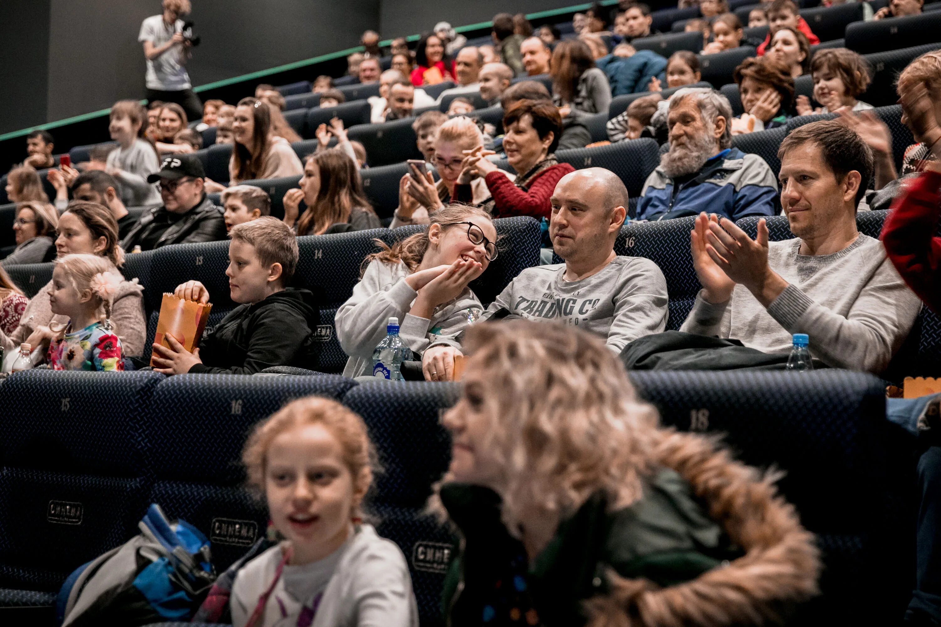 Кинофильмы прокат. Кинотеатр полный зал. Люди в кинотеатре. Много людей в кинотеатре. Очередь в кинотеатр.