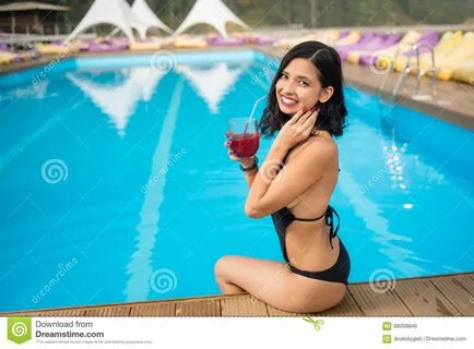Девушка портрета счастливая держа коктеиль, усмехаясь и сидя на краю бассей...